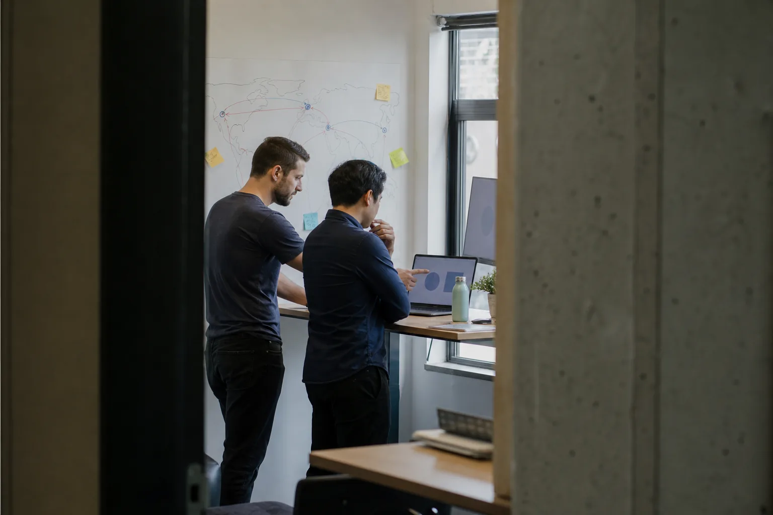 Two developers at a standing desk comparing server regions on a laptop with a world map sketched on a whiteboard behind