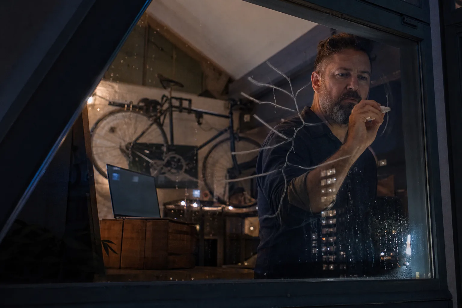 Developer sketching branch lines on a window with a grease pencil in a cramped loft workspace at night, laptop and bike in the background