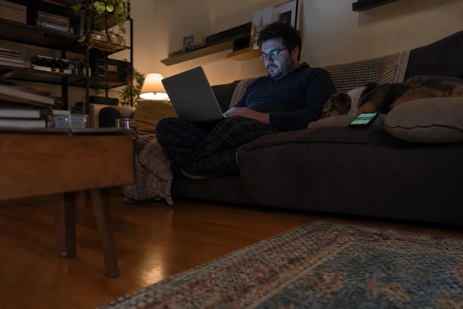 On-call engineer on a sofa at night checking a release on a laptop with a phone showing an alert beside