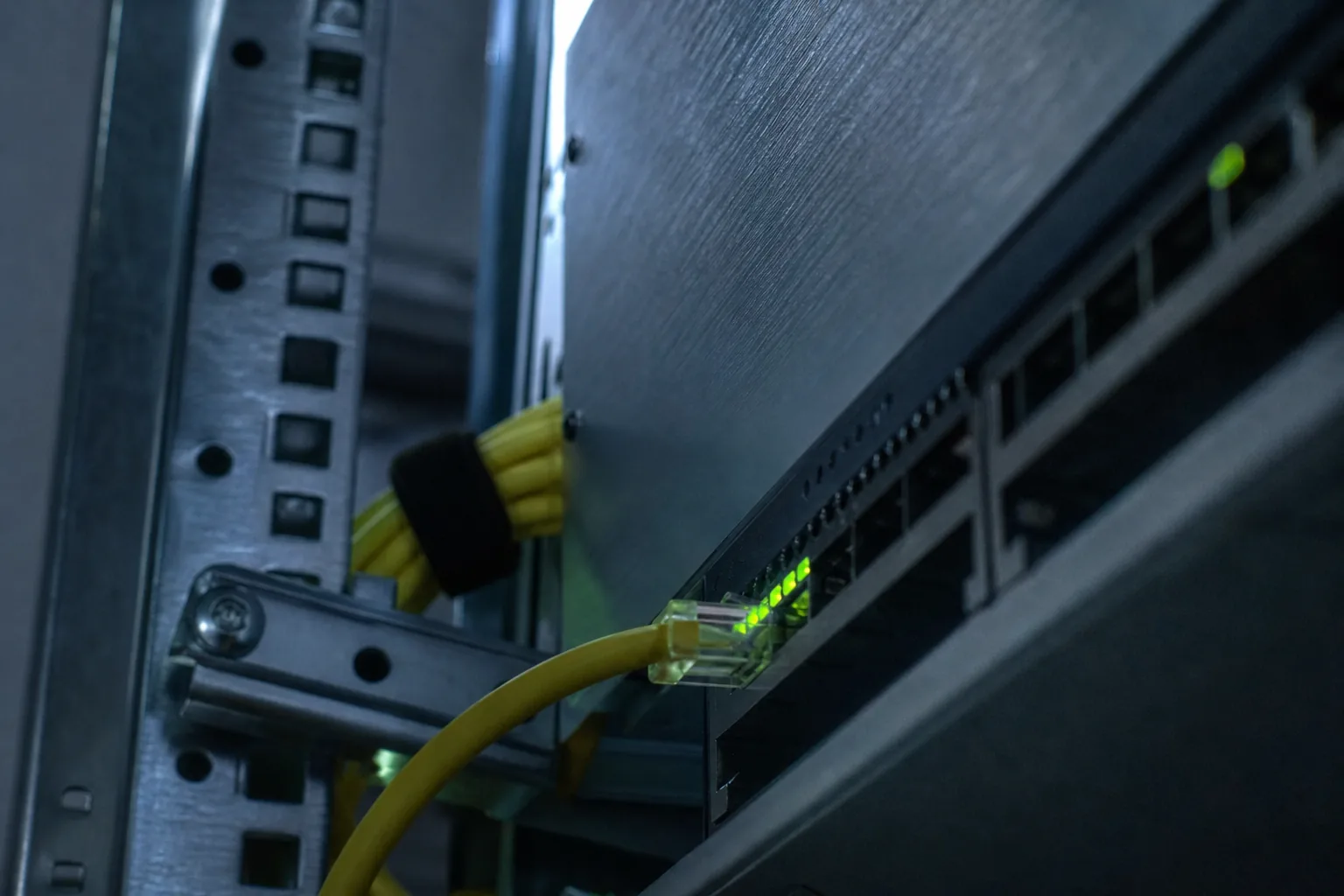 Close-up inside a server rack: a patch cable plugged into a switch port next to a row of small green link lights, out-of-focus cabinet behind